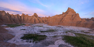 A series of spires lit by the setting sun. Soft pastel colors exist within the sky while grass spreads into the muddy earth. 