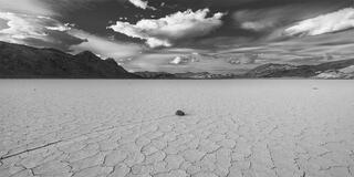 A black and white image of a stone with a mud track behind it as it moves towards the center of the image. Dramatic clouds behind it. 