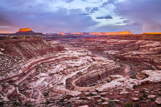 A large red, brown and white canyon cuts through the image with large mesa's lit up by evening light in the distance. 