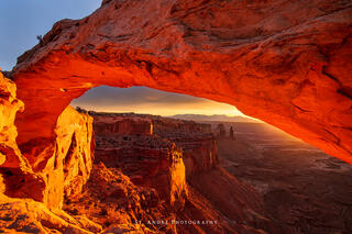 Mesa Arch photographed from the side. Image is deep red with light striking the underside of the arch. Distant mountains and towers can be seen in the distance.