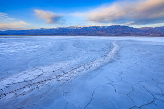 Salt bed with a dried stream leading the viewer into the distance revealing Telescope Peak lit up by the rising sun. The setting moon is somewhat obscured. 