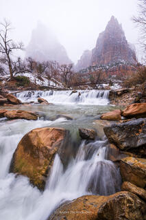 The virgin river flowing over a series of cascades with two towers of the court of the patriarchs are shrouded in mist and snow. 