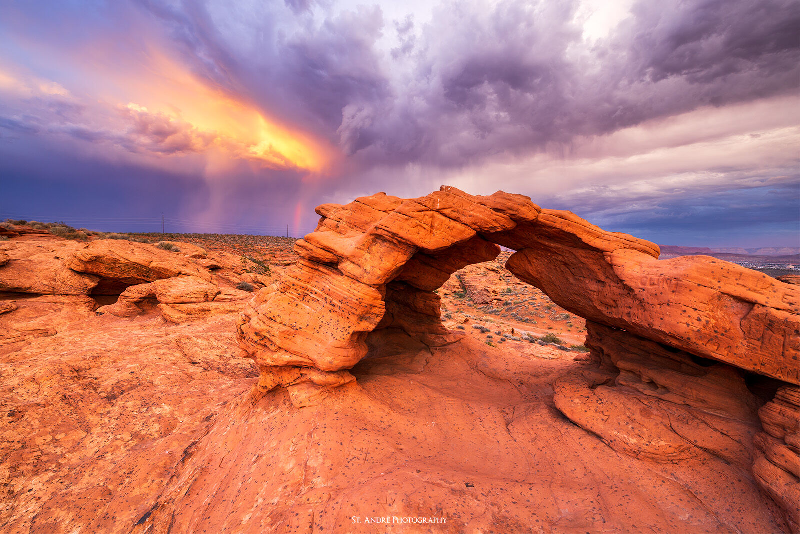 Eagle Arch highlights a desert landscape while a thunderstorm rages above the landscape and a rainbow can be faintly seen to the side of the arch. 
