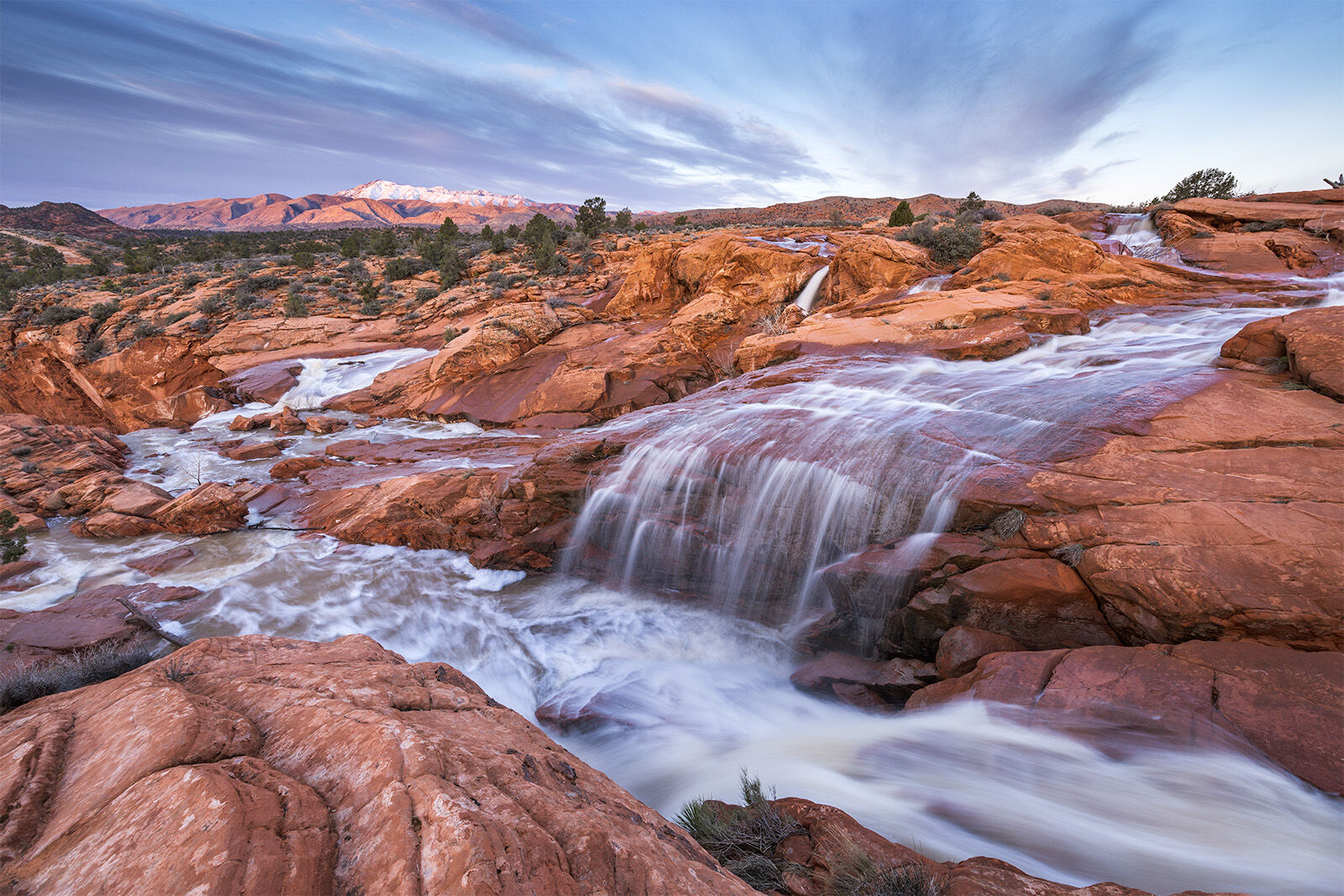 Waterfalls cascading over red rock at sunrise with a distant snow capped mountain in the distance. 
