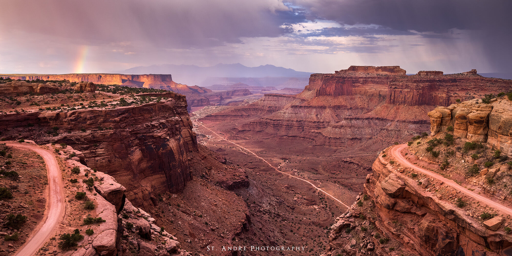 A dirt road winds down through canyons and cliffs with a large rainbow over the cliffs in the distance. 