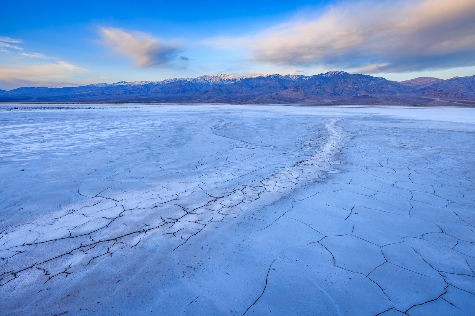 Salt bed with a dried stream leading the viewer into the distance revealing Telescope Peak lit up by the rising sun. The setting moon is somewhat obscured.