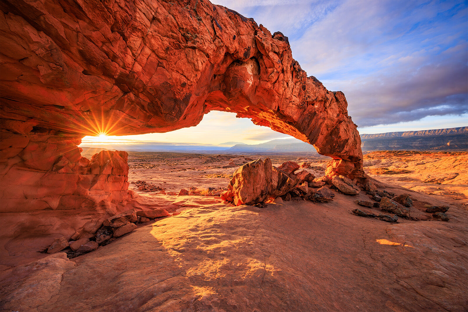 An arch with the sun rising through the corner. The arch is underlit with light and epic clouds swirl in the distance.