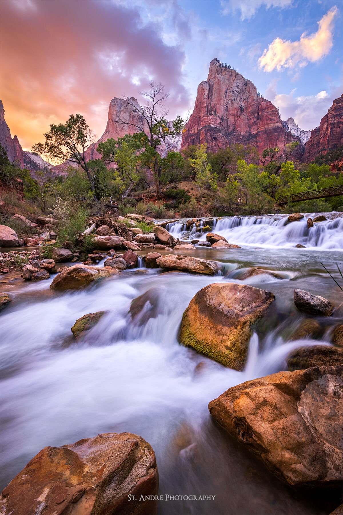 The virgin river cascades down over a series of rocks while two towering mountains reside in the background with sunset lit up clouds.