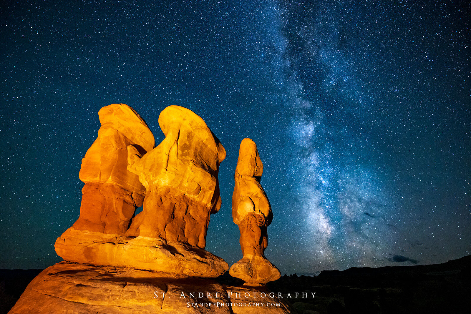 Three large hoodoos lit up by soft warm light with the Milkyway galaxy stretching across the night sky.