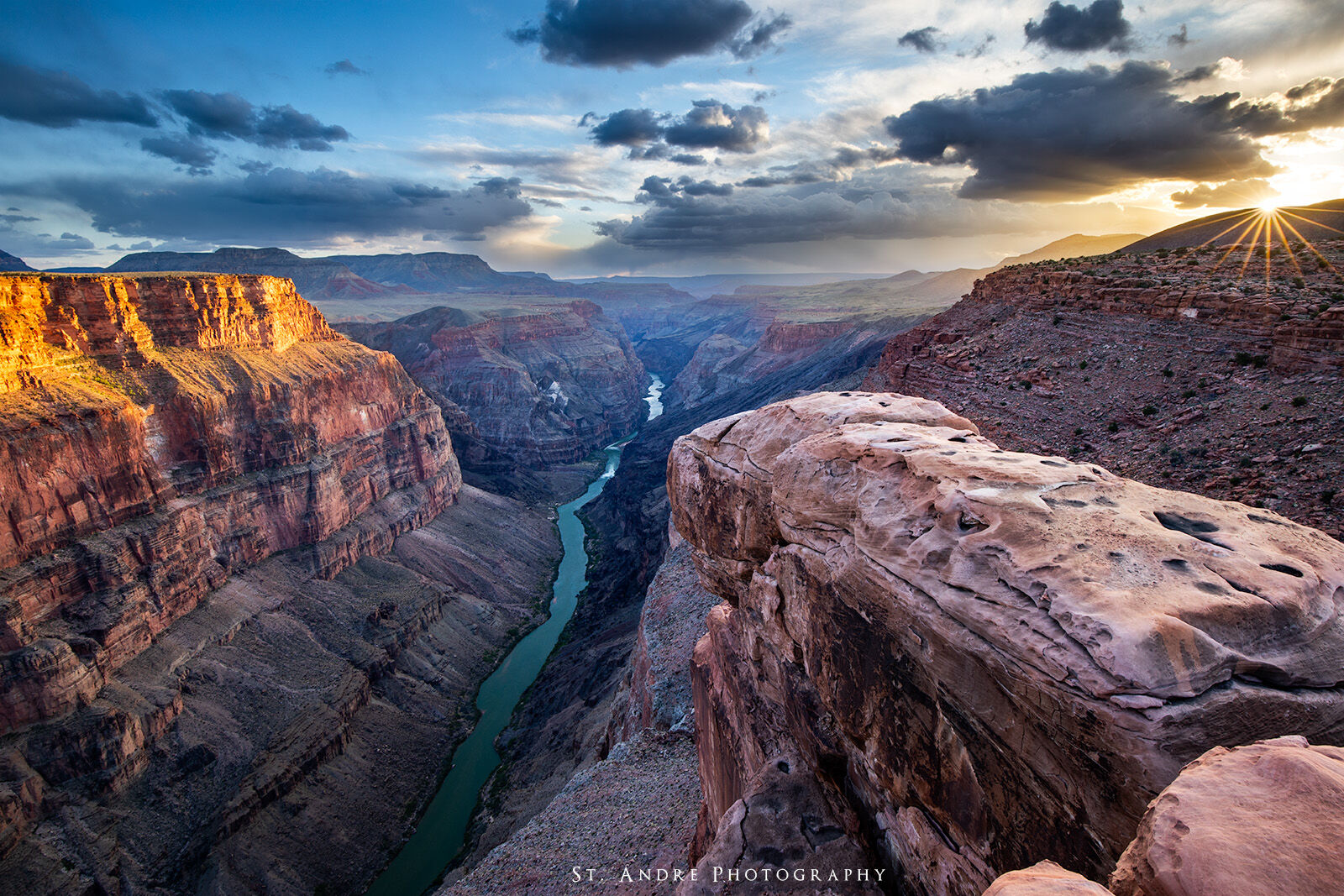 The colorado river cuts through the grand canyon. Large cliffs are on either side of the river. Storm clouds are above and the sun peaks through on the side.  